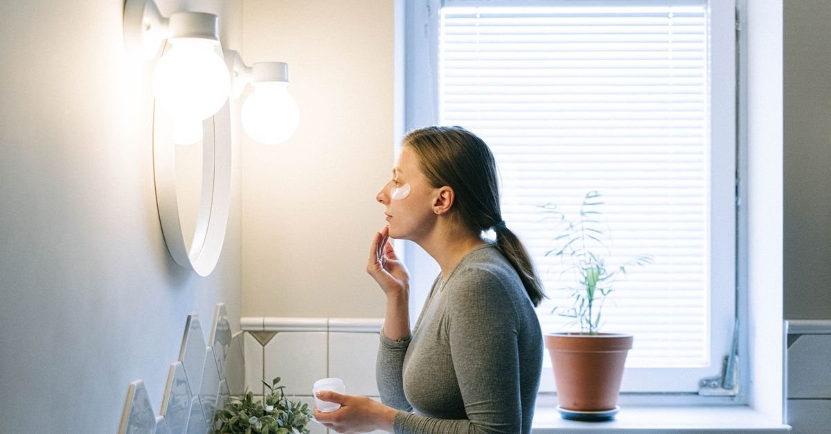 A woman applying face cream by a bright bathroom window