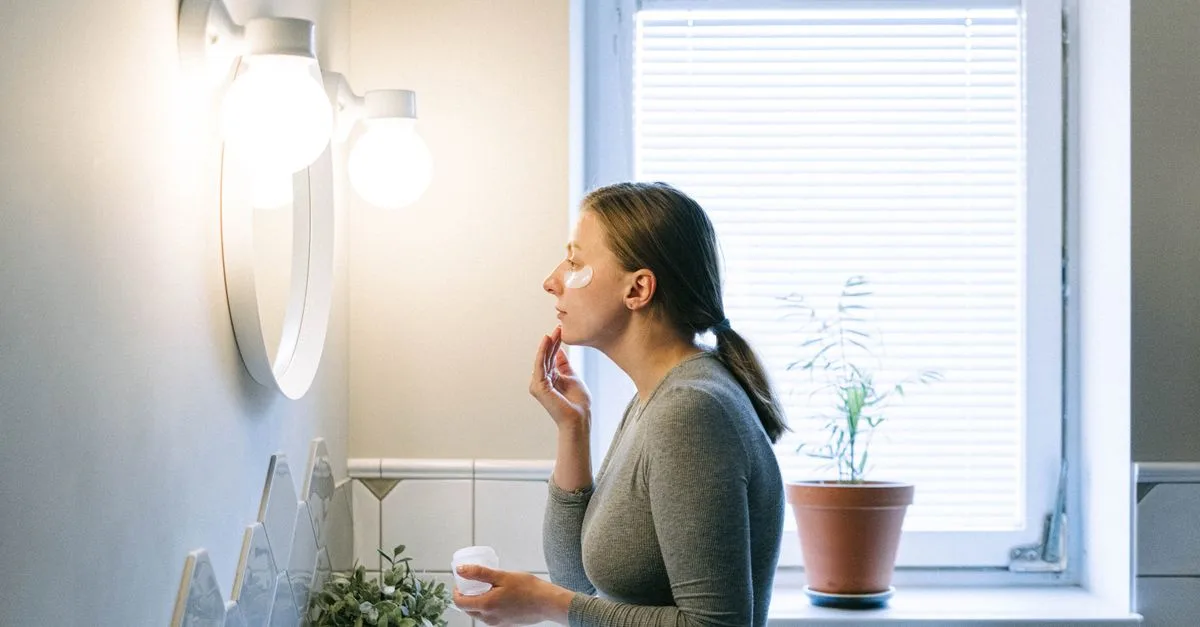 A woman applying face cream by a bright bathroom window