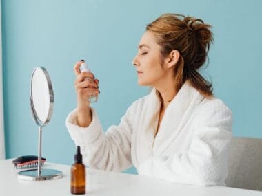 A woman spritzing a facial mist at a vanity mirror