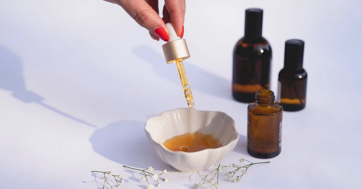 A hand dropping serum into a small bowl beside amber bottles
