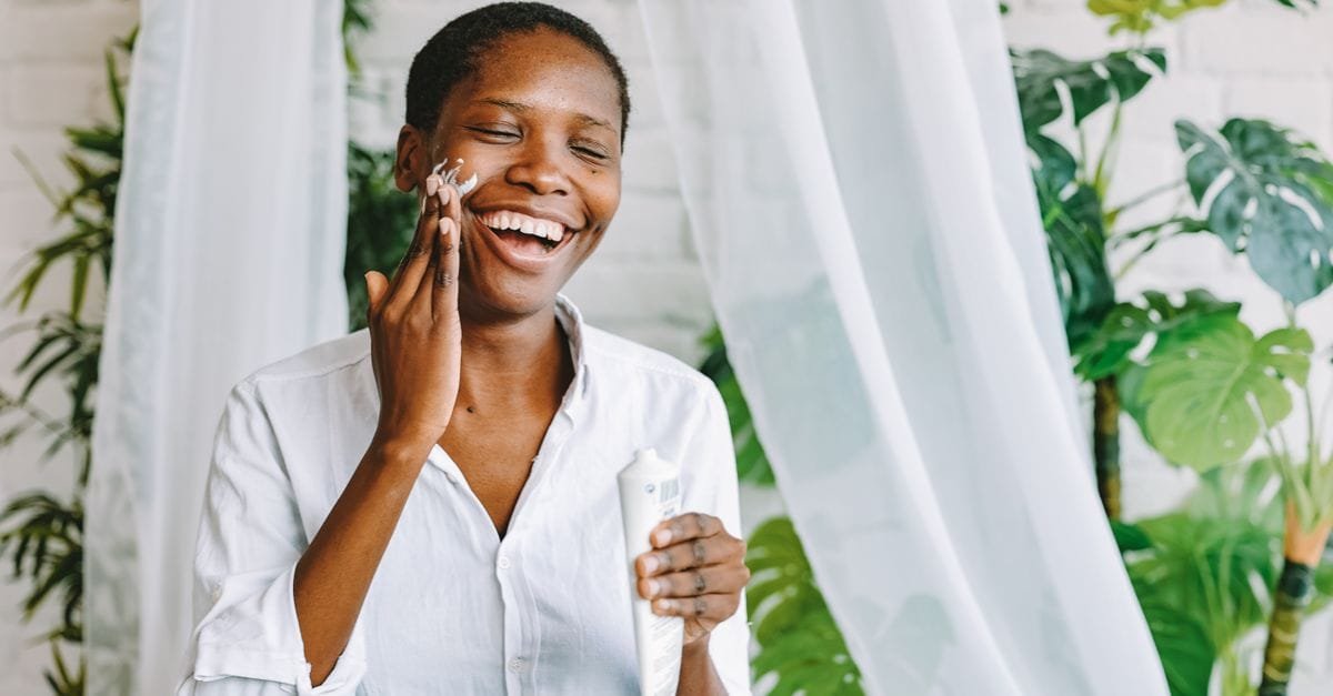 A laughing woman applying face cream by a window