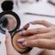 A hand opening a pressed powder compact over a makeup table