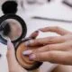 A hand opening a pressed powder compact over a makeup table