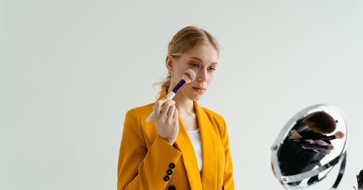 A woman applying makeup at a mirror in a mustard blazer