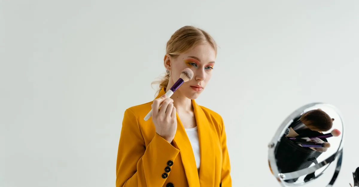 A woman applying makeup at a mirror in a mustard blazer
